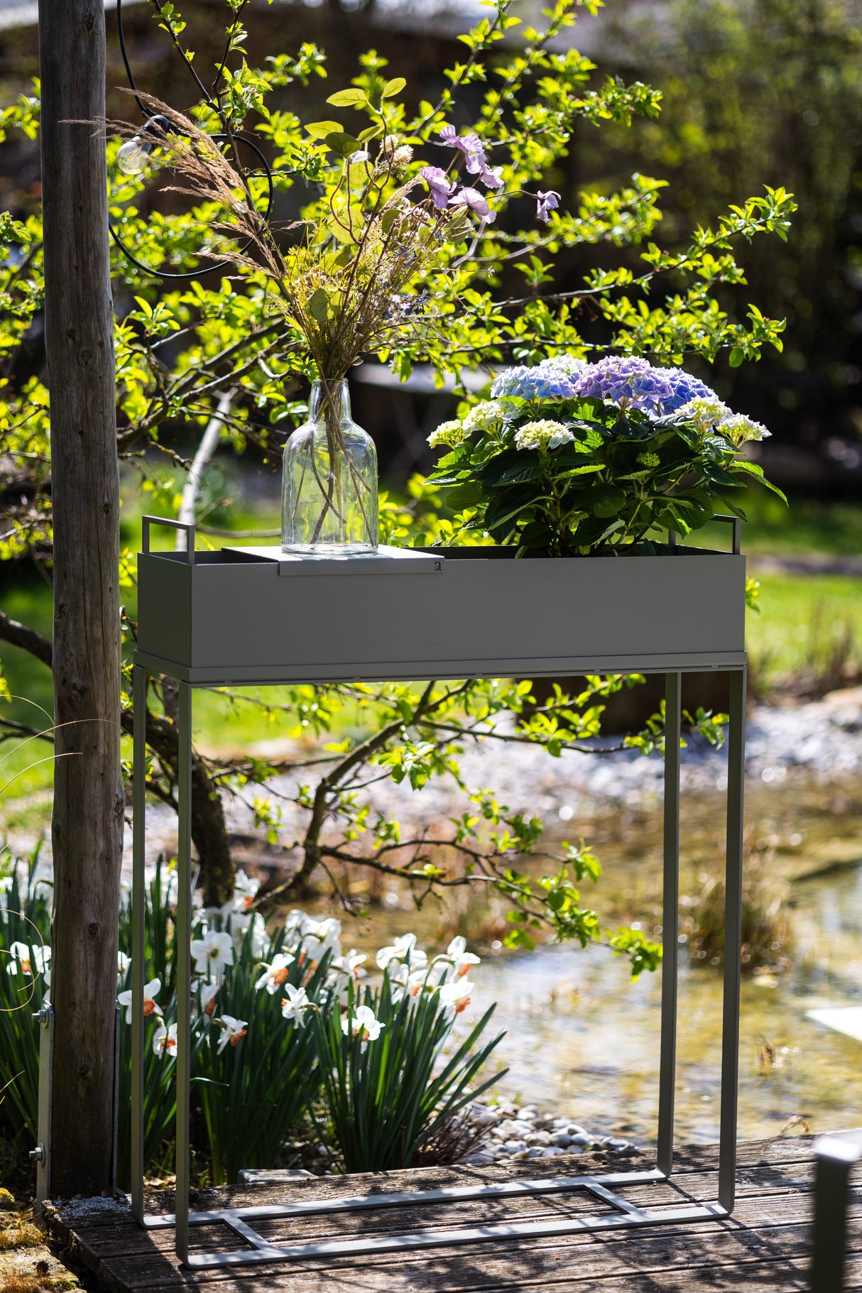 CUBO - Pflanzbehälter auf einem hohen Gestell im Garten, bepflanzt mit Blumen und dekoriert mit einer Glasvase, perfekt für Terrasse oder Balkon.