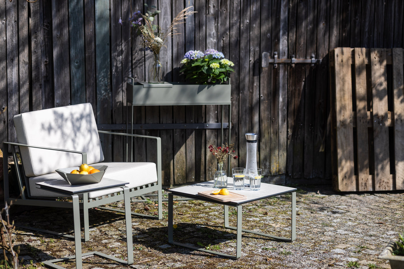 Moderne Gartensitzgruppe mit hellen Polstern auf Steinboden, kleiner Tisch mit Karaffe, Gläsern und Obstschale, im Hintergrund Holzwand mit Pflanzen und Dekoration.