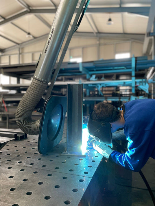 Ein Schweißer mit Schutzmaske arbeitet an einem Metallbauteil in einer Werkstatt mit Absauganlage und professioneller Ausrüstung.