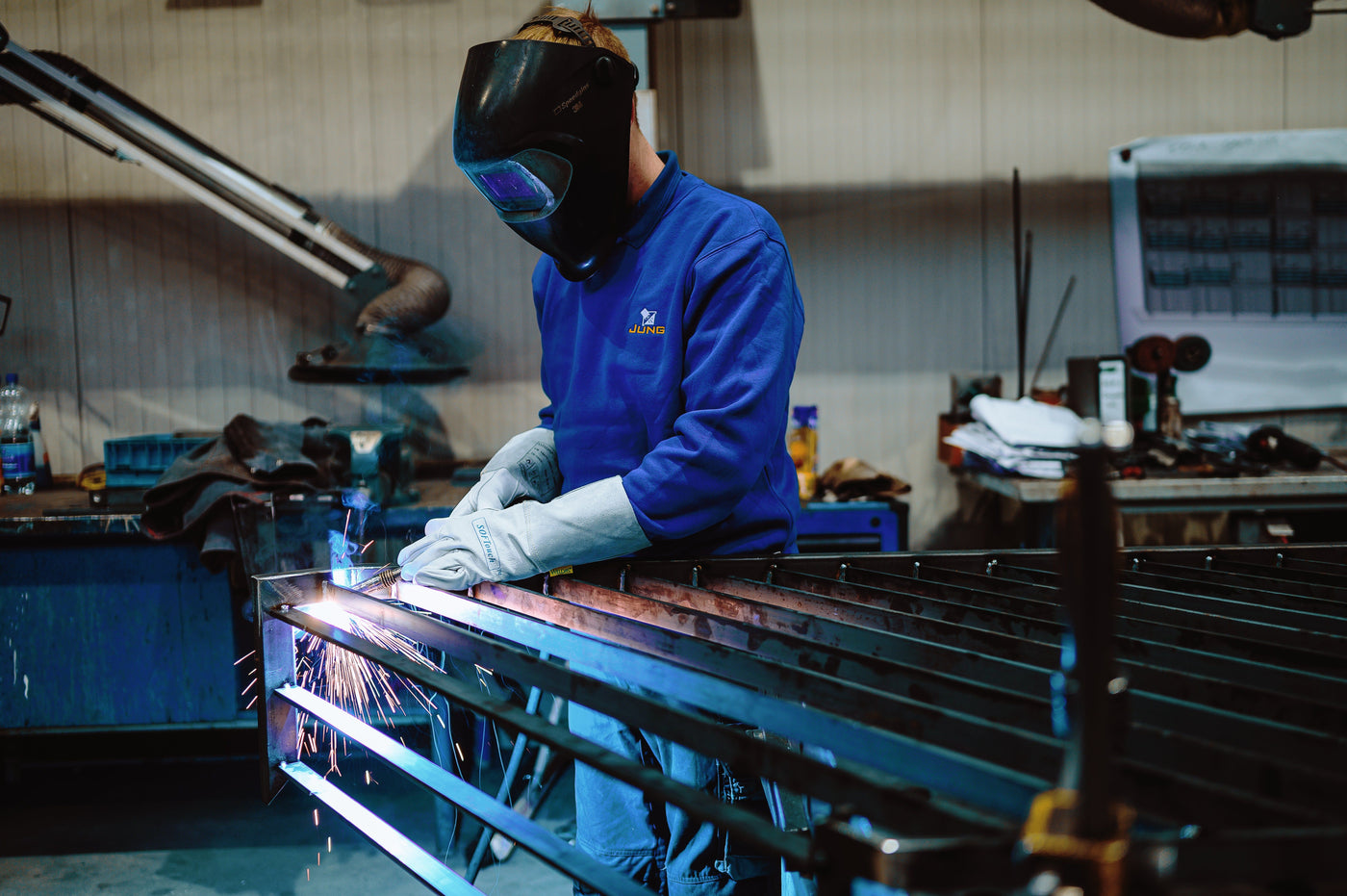 Schweißer mit Schutzhelm und Handschuhen arbeitet mit Lichtbogen-Schweißgerät an Metallkonstruktion in Werkstatt, Funken fliegen beim Schweißen.