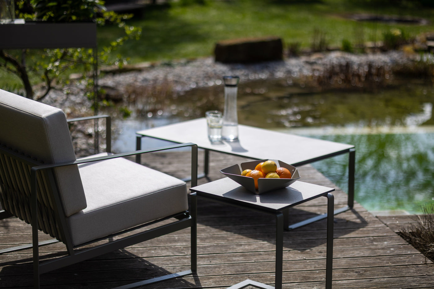 Moderne Terrassenmöbel aus Metall mit hellen Polstern auf einer Holzterrasse am Teich, mit einem Tisch, Karaffe, Glas und einer Schale mit Zitrusfrüchten bei Sonnenschein.